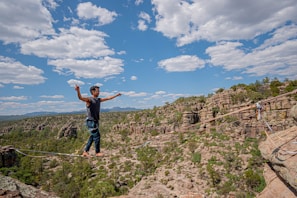 A person is balancing on a tightrope stretched between two rocky cliffs in a mountainous region. The sky is vibrant blue with white clouds scattered across. The landscape features rugged terrain with a mix of green vegetation and rocky outcrops.