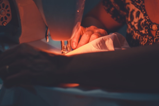 A person is operating a sewing machine, focusing intently as their hands guide a piece of fabric through the machine. The ambiance is warm and cozy, enhanced by soft lighting that illuminates the workspace.