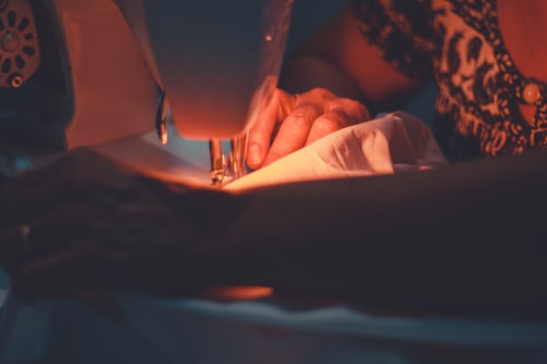 A person is operating a sewing machine, focusing intently as their hands guide a piece of fabric through the machine. The ambiance is warm and cozy, enhanced by soft lighting that illuminates the workspace.