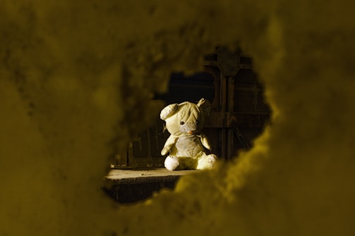 A close-up of a worn-out teddy bear sitting alone on a small bed, bathed in muted grey light.