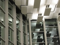 A modern architectural interior with large glass windows and a ceiling featuring rectangular light panels. The design includes a pattern of horizontal and vertical lines, creating a structured and geometric appearance.