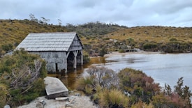 A rustic wooden boathouse sits on the edge of a calm lake surrounded by lush, dense foliage and rolling hills. The structure is slightly weathered, reflecting years of exposure to the elements. The vegetation displays a mix of green and autumnal hues, giving the landscape a natural, untouched feel. The sky is overcast, adding to the tranquil atmosphere.