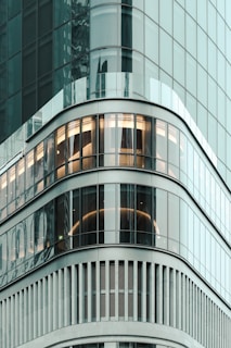 A modern high-rise building with sleek, curved glass windows reflecting the surrounding cityscape. The architectural design features horizontal lines and smooth, rounded edges. Interior lights are visible through the windows, adding warmth to the cool exterior.