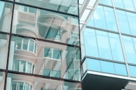 Reflective glass façade of a modern building shows abstract reflections of surrounding structures. The windows create a mirrored effect, including curved and linear architectural details.