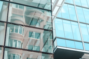 Reflective glass façade of a modern building shows abstract reflections of surrounding structures. The windows create a mirrored effect, including curved and linear architectural details.