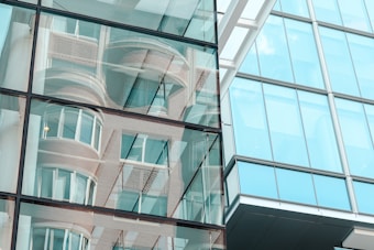 Reflective glass fa&ccedil;ade of a modern building shows abstract reflections of surrounding structures. The windows create a mirrored effect, including curved and linear architectural details.