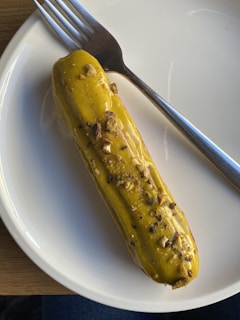 A pickle is placed on a white plate with a fork beside it. The pickle is garnished with chopped nuts or seeds.