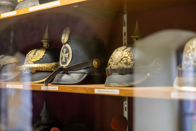 A collection of historical military helmets displayed on wooden shelves. The helmets feature ornate designs with brass and silver decorations, including crest and insignia detailing.