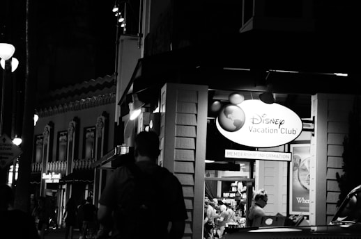 A nighttime street scene featuring a Disney Vacation Club booth with visible signage. The area is populated with people, and there are decorative buildings with lights in the background. The setting appears lively and illuminated by artificial lights.