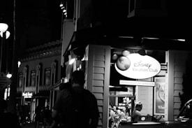 A nighttime street scene featuring a Disney Vacation Club booth with visible signage. The area is populated with people, and there are decorative buildings with lights in the background. The setting appears lively and illuminated by artificial lights.