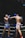 Two Muay Thai fighters are engaged in a match within a boxing ring, each wearing protective gear. One fighter is wearing blue shorts with a determined expression and taking a hit, while the other in pink shorts is delivering a punch with focused intensity. The background includes ropes and advertising banners typical in a sports arena.