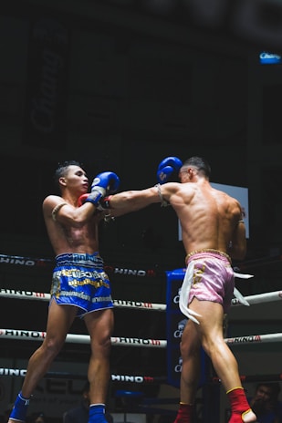Two Muay Thai fighters are engaged in a match within a boxing ring, each wearing protective gear. One fighter is wearing blue shorts with a determined expression and taking a hit, while the other in pink shorts is delivering a punch with focused intensity. The background includes ropes and advertising banners typical in a sports arena.