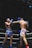 Two Muay Thai fighters are engaged in a match within a boxing ring, each wearing protective gear. One fighter is wearing blue shorts with a determined expression and taking a hit, while the other in pink shorts is delivering a punch with focused intensity. The background includes ropes and advertising banners typical in a sports arena.