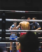 A boxing match is taking place in a ring, with two fighters engaged in combat. One fighter is visible from the front, wearing blue boxing gloves and shorts with Thai writing. The other fighter, seen from the back, is wearing red shorts and gloves. A referee stands nearby, seemingly observing or about to intervene in the match. The environment features dim lighting typical of an indoor sporting event.