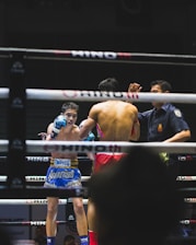 A boxing match is taking place in a ring, with two fighters engaged in combat. One fighter is visible from the front, wearing blue boxing gloves and shorts with Thai writing. The other fighter, seen from the back, is wearing red shorts and gloves. A referee stands nearby, seemingly observing or about to intervene in the match. The environment features dim lighting typical of an indoor sporting event.