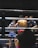 A boxing match is taking place in a ring, with two fighters engaged in combat. One fighter is visible from the front, wearing blue boxing gloves and shorts with Thai writing. The other fighter, seen from the back, is wearing red shorts and gloves. A referee stands nearby, seemingly observing or about to intervene in the match. The environment features dim lighting typical of an indoor sporting event.
