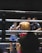 A boxing match is taking place in a ring, with two fighters engaged in combat. One fighter is visible from the front, wearing blue boxing gloves and shorts with Thai writing. The other fighter, seen from the back, is wearing red shorts and gloves. A referee stands nearby, seemingly observing or about to intervene in the match. The environment features dim lighting typical of an indoor sporting event.