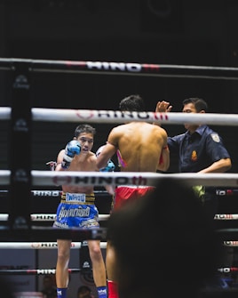 A boxing match is taking place in a ring, with two fighters engaged in combat. One fighter is visible from the front, wearing blue boxing gloves and shorts with Thai writing. The other fighter, seen from the back, is wearing red shorts and gloves. A referee stands nearby, seemingly observing or about to intervene in the match. The environment features dim lighting typical of an indoor sporting event.