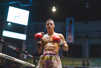 A muscular boxer stands confidently in a boxing ring, wearing red gloves and colorful shorts under bright arena lights. The dimly lit background shows a large screen and banners hanging from the ceiling.