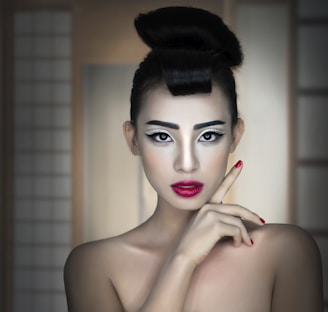 A woman with an elaborate hairstyle and dramatic makeup, including bold eyebrows, heavy eyeliner, and bright red lipstick. She is looking directly at the camera with a poised expression, one hand raised near her face.