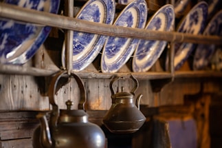 An arrangement of blue and white decorative plates is displayed on a wooden rack. Below the plates, there is an antique metal kettle with a curved spout and handle, hanging from the wood. The setting evokes a rustic and traditional atmosphere with a focus on the intricate design of the plates and the aged look of the metal kettle.