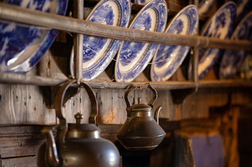 An arrangement of blue and white decorative plates is displayed on a wooden rack. Below the plates, there is an antique metal kettle with a curved spout and handle, hanging from the wood. The setting evokes a rustic and traditional atmosphere with a focus on the intricate design of the plates and the aged look of the metal kettle.
