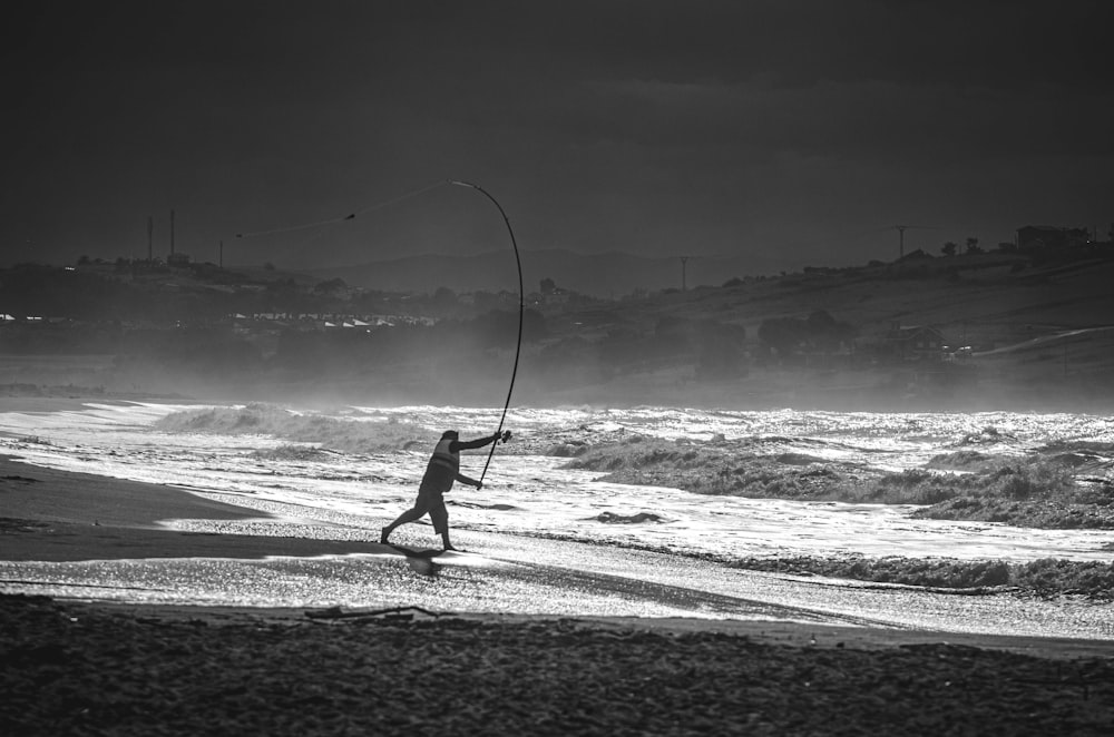grayscale photo of person holding fishing rod walking on beach shore