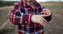 A child wearing a plaid red shirt plays with a grasshopper perched on their arm, with an outdoor background of grass and trees.