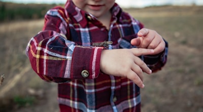 A child wearing a plaid red shirt plays with a grasshopper perched on their arm, with an outdoor background of grass and trees.