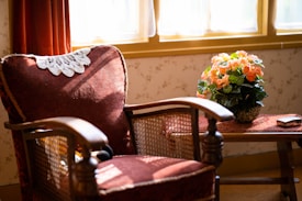 A vintage armchair with a lace doily draped over the backrest is positioned by a window, bathed in warm sunlight. Beside the chair, on a small wooden table, sits a pot of vibrant orange flowers with lush green leaves. The decor suggests a cozy, inviting atmosphere with a touch of nostalgia.
