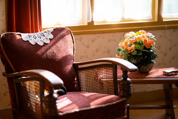 A vintage armchair with a lace doily draped over the backrest is positioned by a window, bathed in warm sunlight. Beside the chair, on a small wooden table, sits a pot of vibrant orange flowers with lush green leaves. The decor suggests a cozy, inviting atmosphere with a touch of nostalgia.