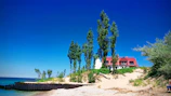 A beach scene features a house with a red roof and a white lighthouse behind a row of tall, narrow trees. The beach is sandy with some vegetation and a clear blue sky above. The ocean is visible on the left side, and part of an old wooden structure is seen along the shoreline.