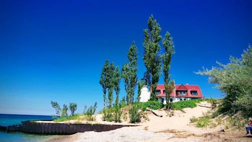 A beach scene features a house with a red roof and a white lighthouse behind a row of tall, narrow trees. The beach is sandy with some vegetation and a clear blue sky above. The ocean is visible on the left side, and part of an old wooden structure is seen along the shoreline.