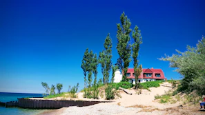 A beach scene features a house with a red roof and a white lighthouse behind a row of tall, narrow trees. The beach is sandy with some vegetation and a clear blue sky above. The ocean is visible on the left side, and part of an old wooden structure is seen along the shoreline.