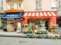 A street scene featuring a charming neighborhood with a boucherie on the left and a flower shop on the right. The boucherie has a navy blue awning with lettering, displaying a variety of meats. The flower shop, with a red and white striped awning, showcases a vibrant assortment of flowers, attracting a passerby who is browsing the colorful display.