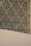 Intricate lattice structure forms a geometric pattern on a modern building facade. The design consists of intersecting diagonal metal bars creating diamond-shaped segments. The lower portion of the image shows a large, flat concrete surface contrasting with the complex design above.