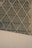 Intricate lattice structure forms a geometric pattern on a modern building facade. The design consists of intersecting diagonal metal bars creating diamond-shaped segments. The lower portion of the image shows a large, flat concrete surface contrasting with the complex design above.
