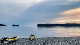 Two kayaks are resting on a pebbled beach, with calm water and a distant view of tree-covered islands. The sky is overcast with soft hues of blue and orange from the setting or rising sun.