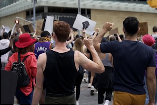 A crowd of people participate in a protest, holding up signs and raising their fists. The individuals are diverse in appearance, wearing casual clothing, and one person is notably wearing a bright red hat. The atmosphere conveys a sense of unity and determination.