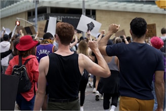 A crowd of people participate in a protest, holding up signs and raising their fists. The individuals are diverse in appearance, wearing casual clothing, and one person is notably wearing a bright red hat. The atmosphere conveys a sense of unity and determination.