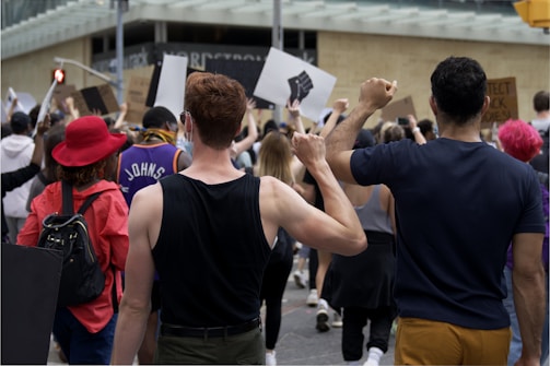 A crowd of people participate in a protest, holding up signs and raising their fists. The individuals are diverse in appearance, wearing casual clothing, and one person is notably wearing a bright red hat. The atmosphere conveys a sense of unity and determination.