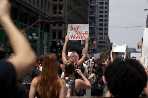 A group of people participate in a protest on a city street, holding up signs. Many signs are unreadable except for one clearly stating 'Say Their Names'. The protestors appear diverse, and their expressions suggest determination and solidarity. Towering buildings and traffic lights form the urban backdrop.