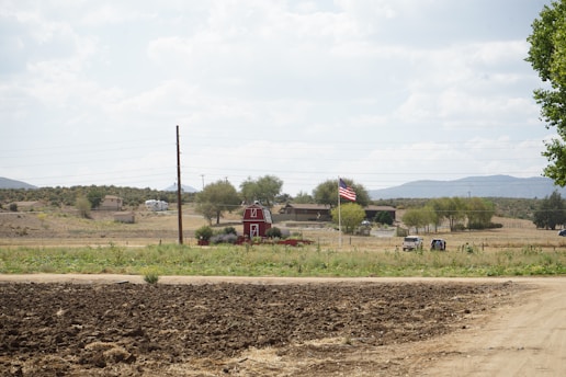 A rural landscape featuring a red barn with a gambrel roof situated amid a grassy field. A dirt path runs across the foreground, and an American flag is prominently displayed nearby. Trees and distant hills frame the horizon, with houses and vehicles visible in the background.