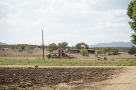 A rural landscape featuring a red barn with a gambrel roof situated amid a grassy field. A dirt path runs across the foreground, and an American flag is prominently displayed nearby. Trees and distant hills frame the horizon, with houses and vehicles visible in the background.