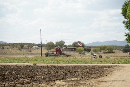 A rural landscape featuring a red barn with a gambrel roof situated amid a grassy field. A dirt path runs across the foreground, and an American flag is prominently displayed nearby. Trees and distant hills frame the horizon, with houses and vehicles visible in the background.