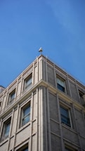 A corner of a multi-story building with a classic brick facade under a clear blue sky. The building features large windows with decorative moldings and a security camera mounted at the top corner, facing outward.