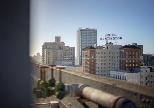 A view of several mid-rise buildings with a prominent sign for Hotel Huntington on top of one of the structures. The scene is taken from a nearby rooftop, partially showing industrial pipes with red valve handles in the foreground. The lighting suggests it is late afternoon, casting soft shadows on the buildings.
