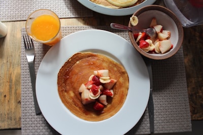 A breakfast scene featuring a plate with a crepe topped with sliced strawberries and bananas. Off to the side, there is a bowl with additional fruit slices and a spoon. Next to the plate is a glass of orange juice. The setting is on a wooden table with a woven placemat, and there is a fork placed beside the plate.