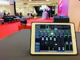 A tablet is displaying audio mixing software on a table, with a large hall visible in the background. The room is set up for an event, with red carpet flooring, seating arrangements, and a stage area where two people stand next to a table with balloons.