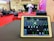 A tablet is displaying audio mixing software on a table, with a large hall visible in the background. The room is set up for an event, with red carpet flooring, seating arrangements, and a stage area where two people stand next to a table with balloons.